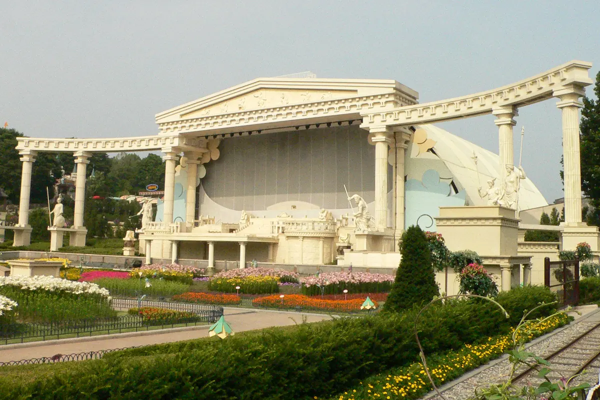 A white open-air stage with Greek-style columns and statues stands behind flower beds and a clipped hedge
